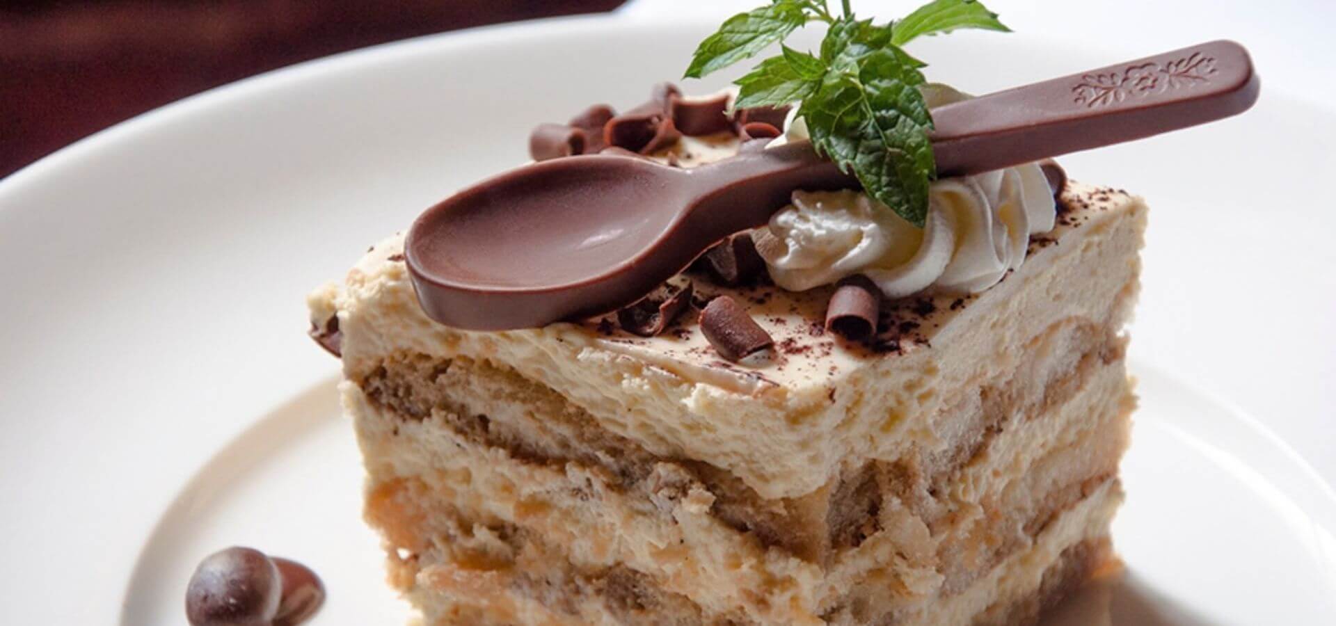 A slice of layered dessert topped with a mint leaf, chocolate curls, and a chocolate spoon.
