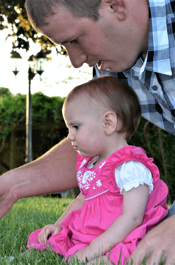 A man leans down to interact with a young girl in a pink dress sitting on the grass.