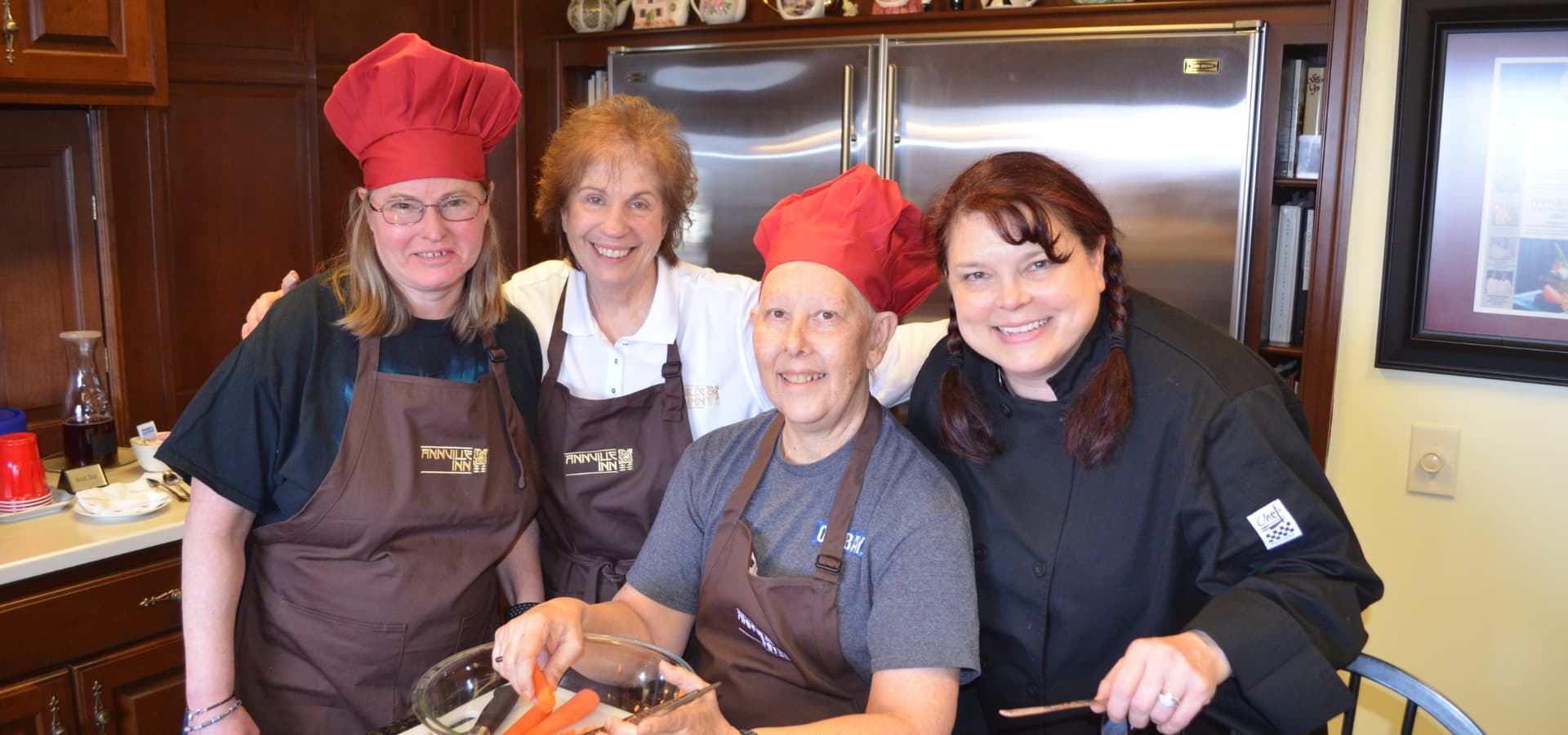 Four women, wearing chef hats and aprons, smile together in a kitchen while preparing food.