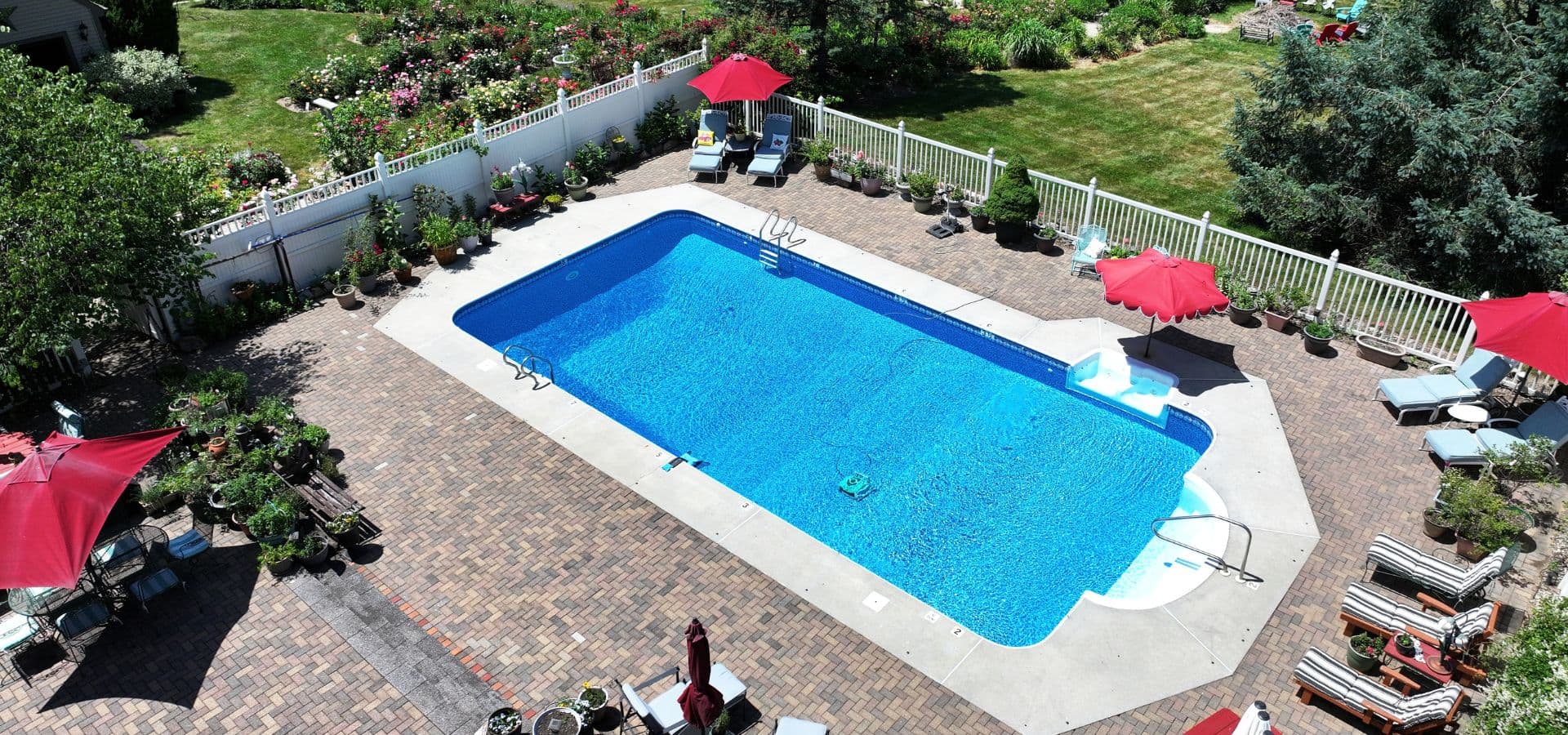 Aerial view of a blue swimming pool surrounded by lounge chairs and red umbrellas in a well-manicured garden.