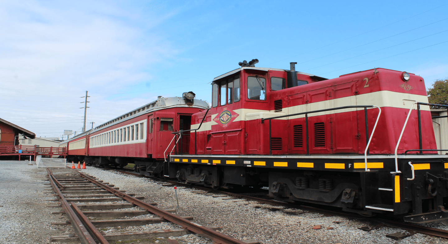 A red and white diesel locomotive is parked next to a vintage passenger car on railway tracks under a blue sky.