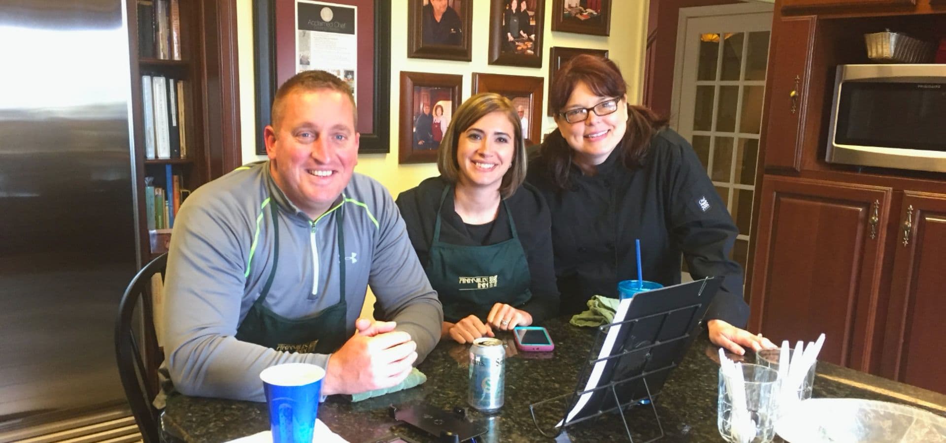 Three people sit at a kitchen counter, smiling and chatting, with framed photos on the wall behind them.