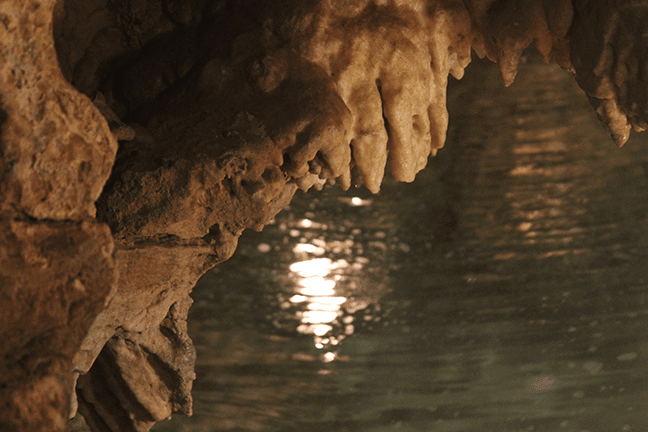 Stalactite-like formations hang above a calm, reflective water surface.