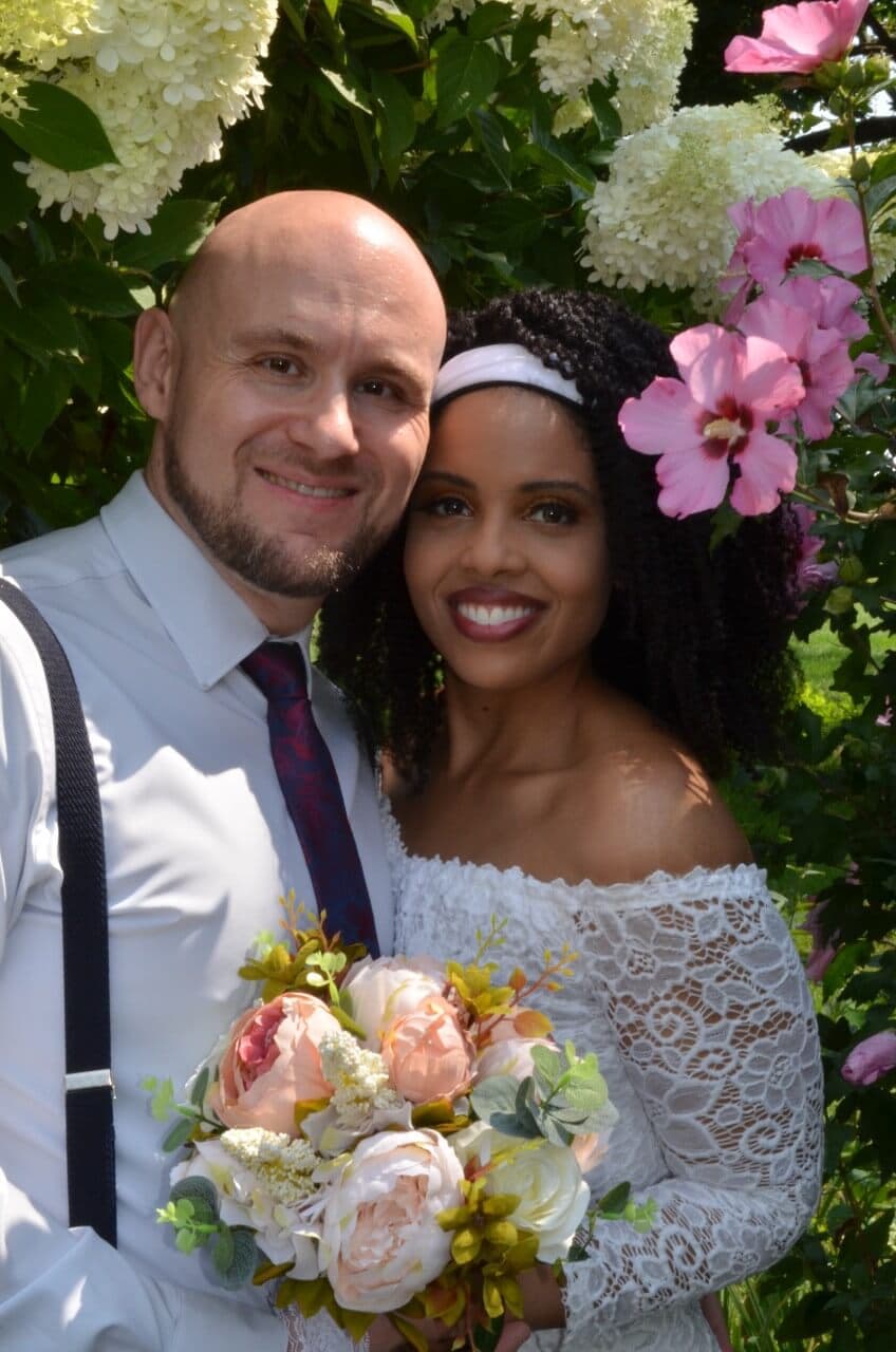 A smiling couple stands together in a garden, surrounded by flowers, with the woman holding a bouquet.