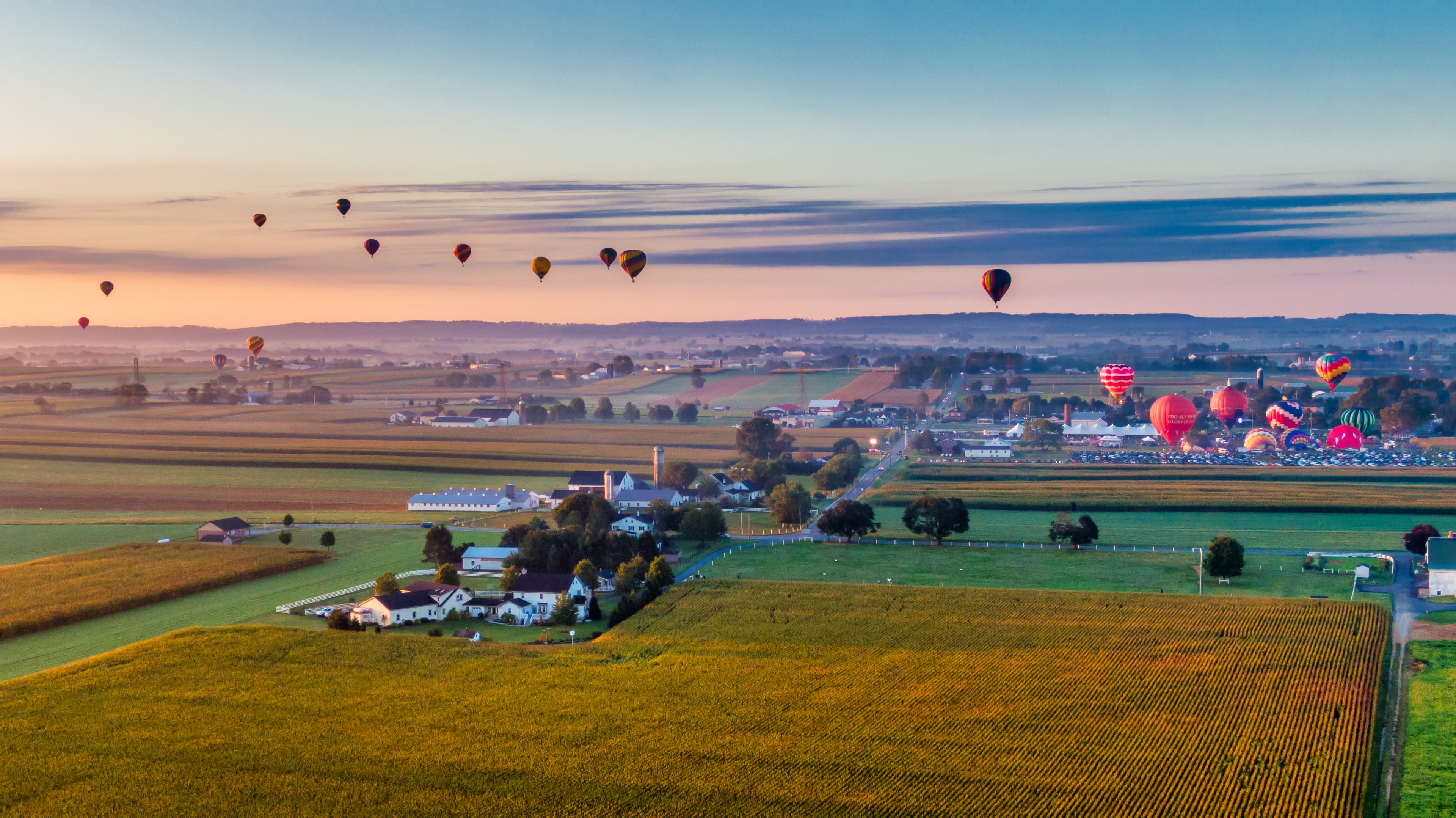 A panoramic view of a countryside landscape with multiple hot air balloons floating above fields at sunrise.