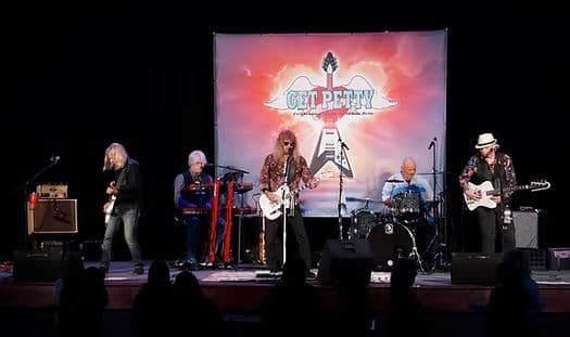A tribute band performs on stage in front of an audience, with a backdrop featuring a guitar logo.