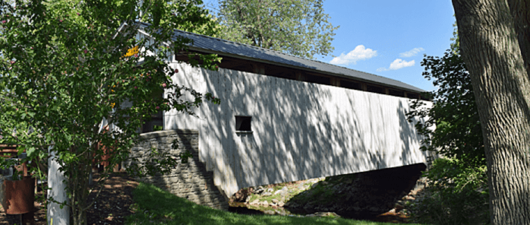 A modern covered bridge nestled among trees, spanning a small creek.