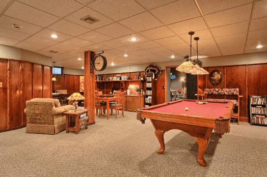 A cozy basement game room featuring a pool table, lounge area, and shelves filled with books and DVDs.