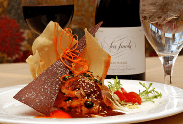 A gourmet dish garnished with colorful vegetables, accompanied by red wine and decorative elements on a fine dining table.