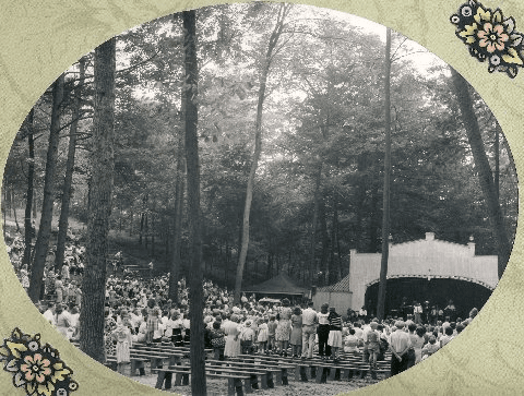 A large crowd gathers in a wooded area to watch a performance on stage.