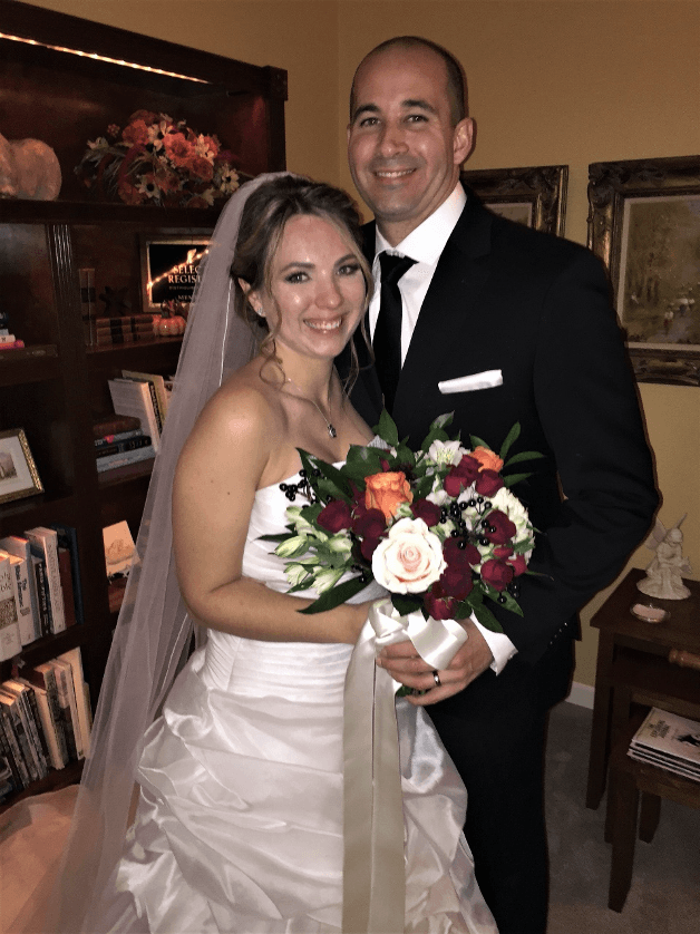 A smiling bride in a white gown holds a bouquet while standing with a groom in a black suit.