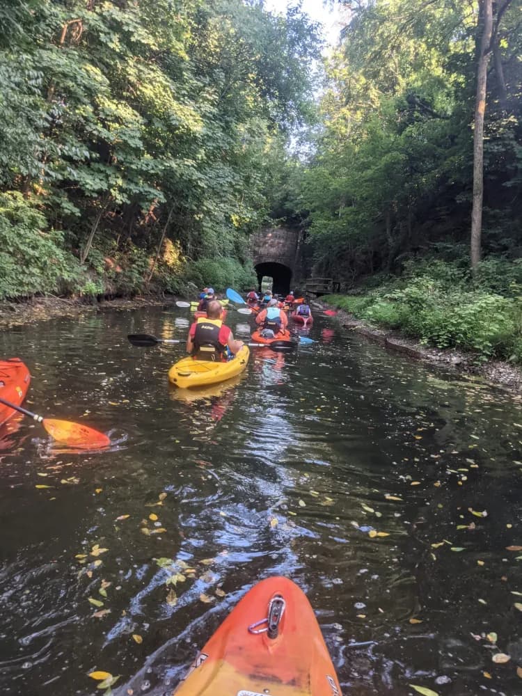 A group of kayakers paddles through a narrow, tree-lined waterway leading to a dark tunnel.