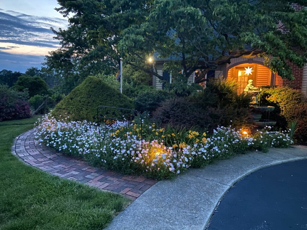 A beautifully landscaped garden with flowers, a brick pathway, and warm evening lighting near a home.