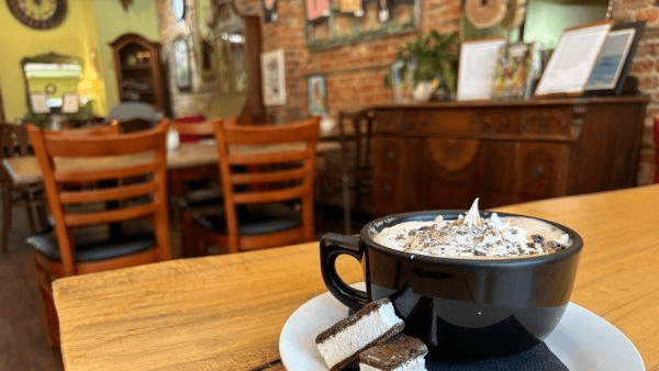 A black coffee cup topped with whipped cream and chocolate sits on a wooden table beside a plate of chocolate dessert, with a cozy café background.