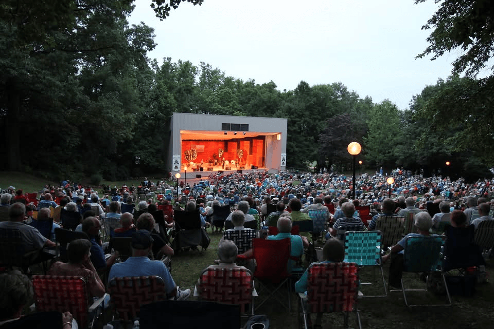 A large crowd of people seated in folding chairs watches a performance on an outdoor stage surrounded by trees in the early evening.