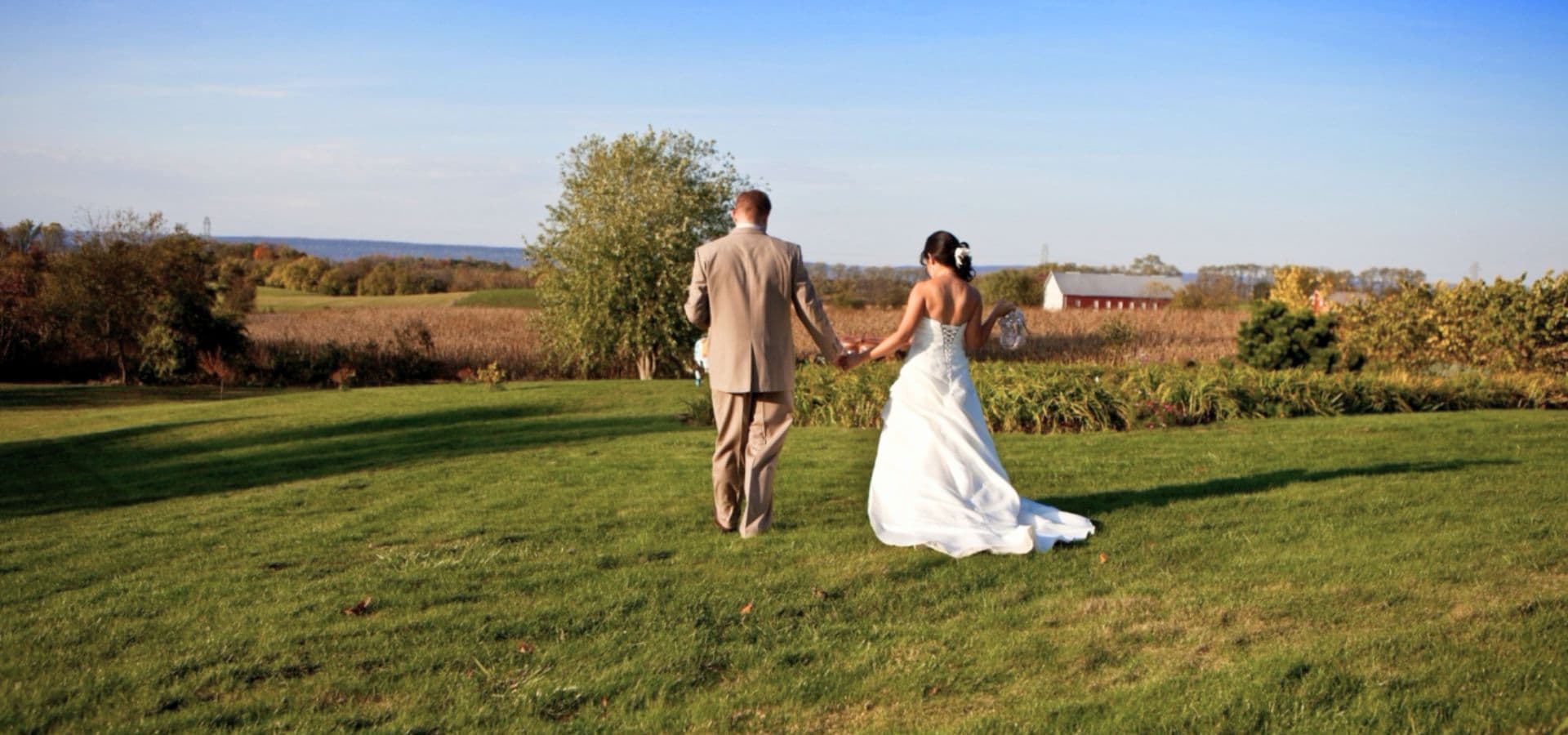 A couple walks hand-in-hand through a lush green field, with a scenic landscape in the background.