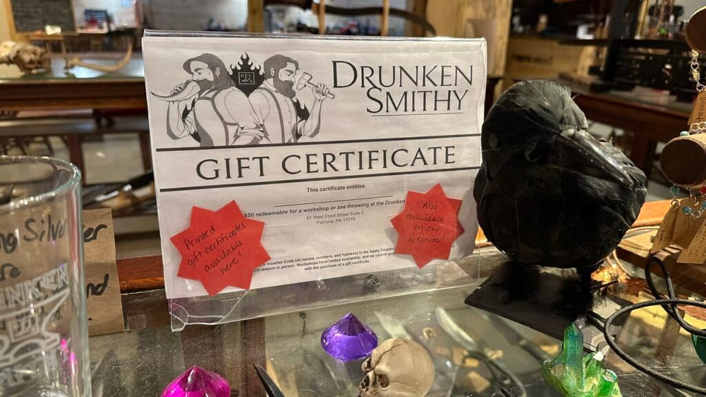 A gift certificate for Drunken Smithy displayed on a countertop with a black crow statue and colorful decorative gems.