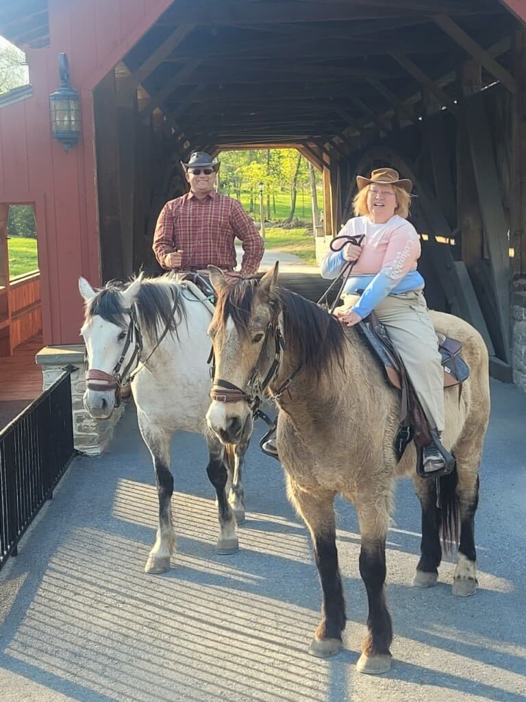 A man and a woman ride horses through a covered bridge on a sunny day.