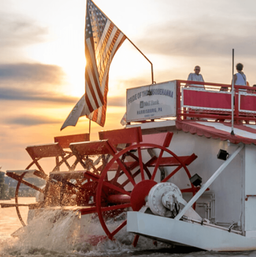 A riverboat with paddle wheels, flying an American flag, cruises at sunset.