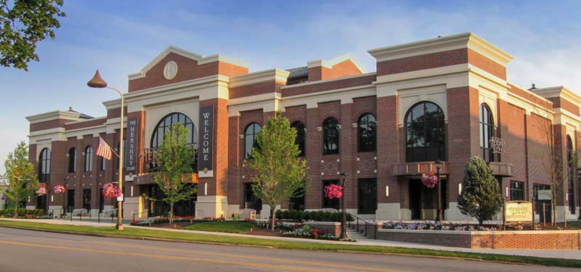 A large brick building with arched windows and a welcoming sign, surrounded by trees and flowers.