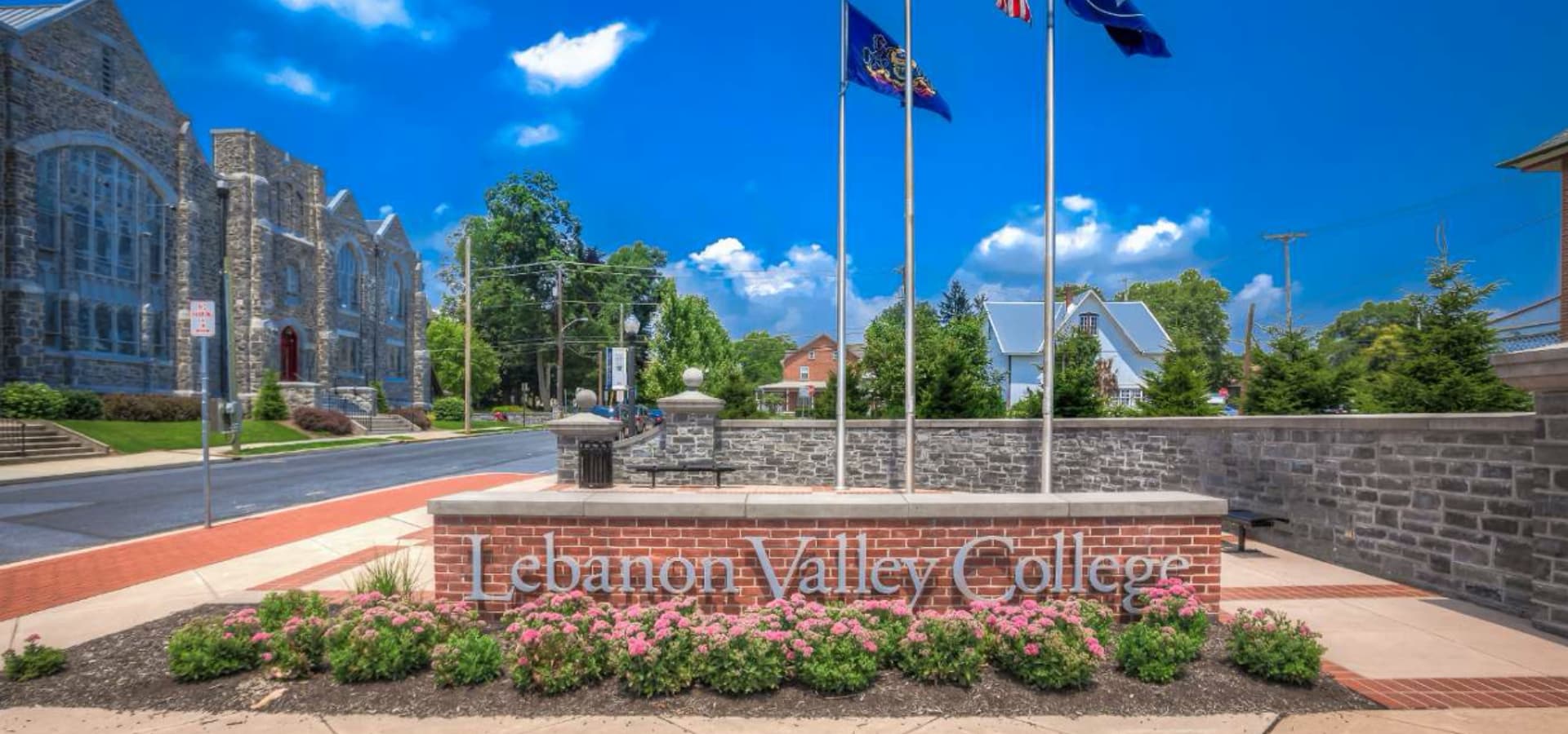 Sign for Lebanon Valley College with flags and greenery in a sunny setting.