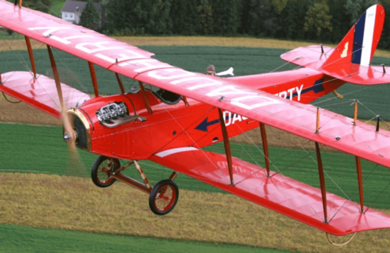 A red biplane flying over green fields.