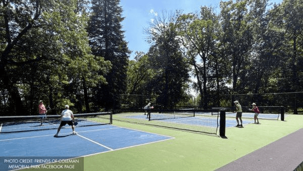 Two tennis courts with players engaged in a match surrounded by greenery on a sunny day.