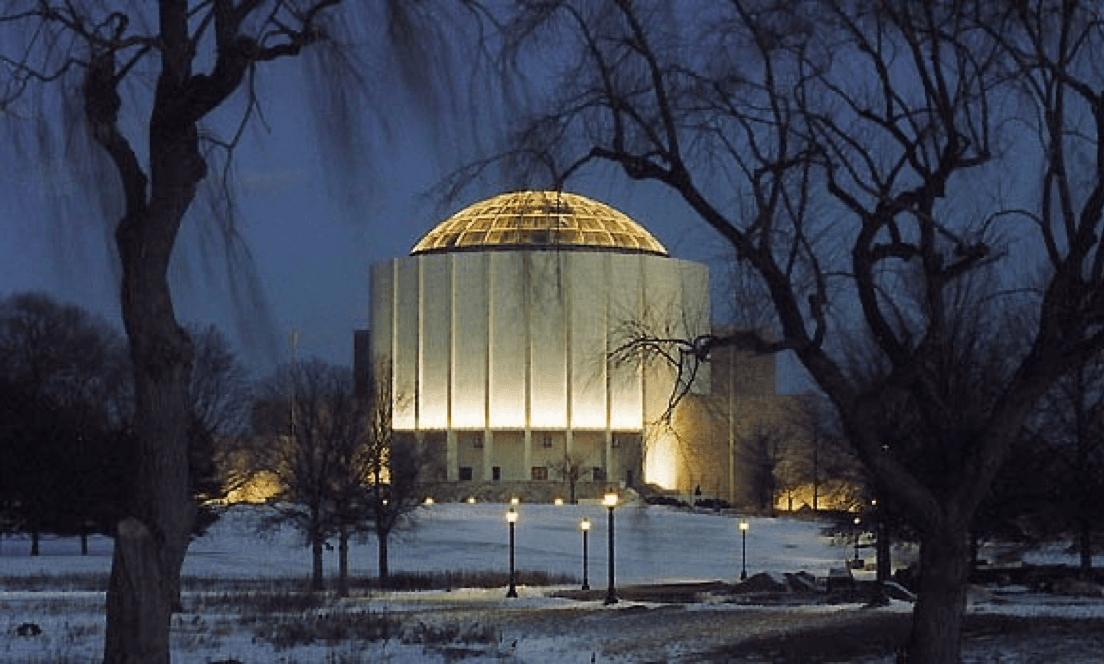A brightly lit dome-shaped building surrounded by bare trees at night.