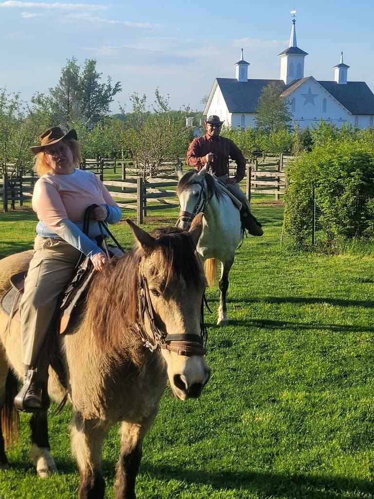 Two riders on horseback in a green field with a white barn in the background.