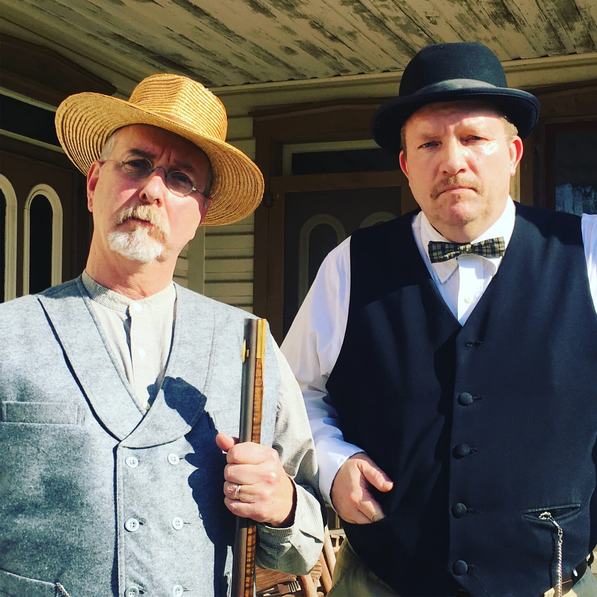 Two men in vintage attire pose together outdoors, one wearing a straw hat and the other a bowler hat.