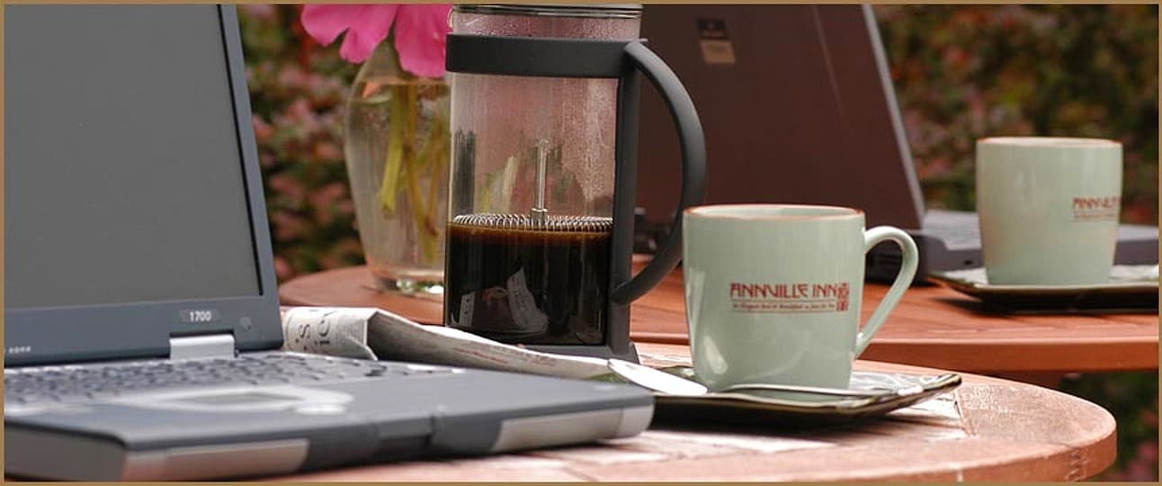 A coffee cup and French press on a table beside two laptops amid a floral background.