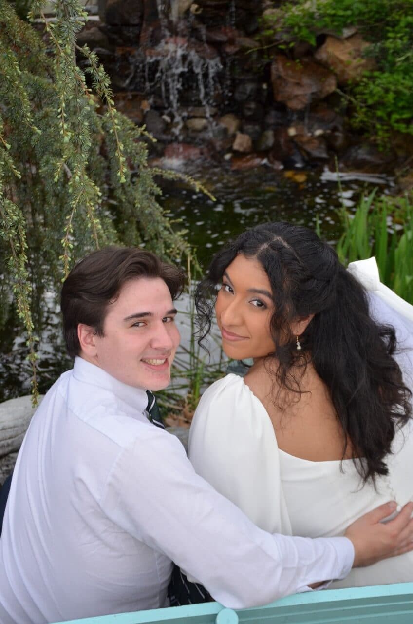 A couple poses together by a serene pond with a waterfall in the background.