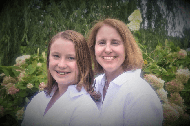 A girl and a woman smile together in matching white shirts, surrounded by greenery and flowers.