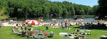 A busy lakeside beach scene with sunbathers and colorful umbrellas on a sunny day.