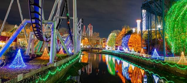 A nighttime view of a theme park featuring colorful holiday lights and roller coaster tracks reflected in a nearby waterway.