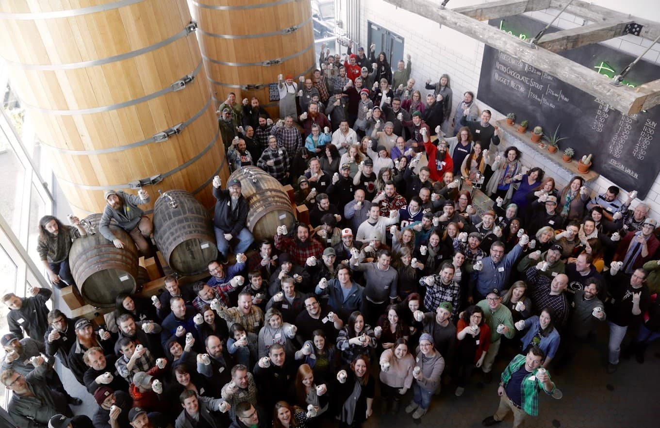 A large group of people gathered indoors, smiling and holding drinks near wooden barrels.