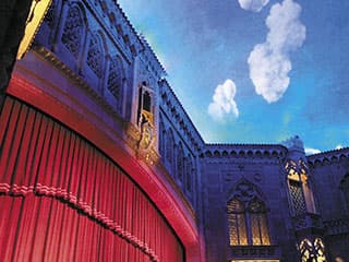 A vibrant theater interior with ornate architecture and a red curtain against a painted sky backdrop.