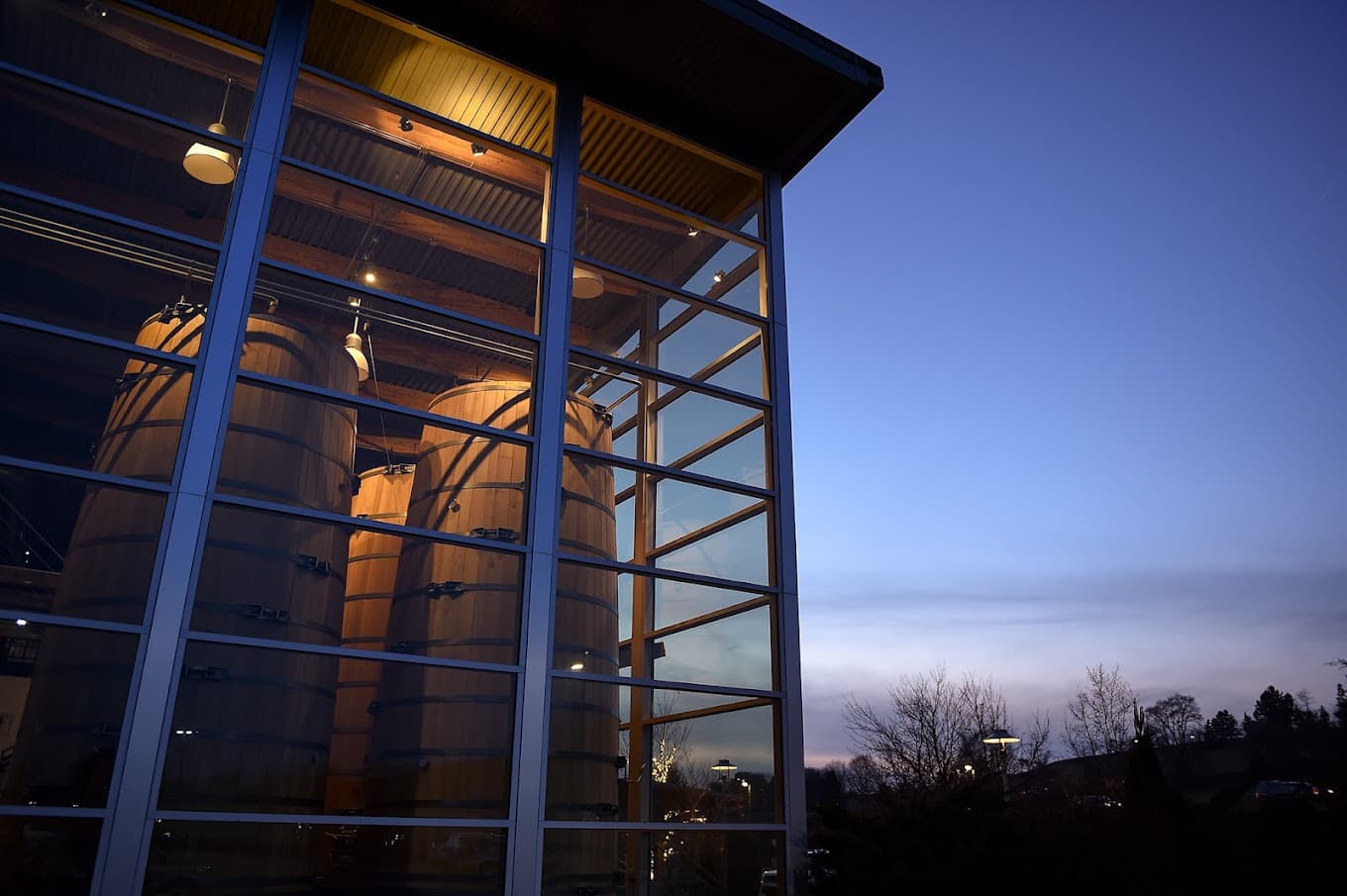 A modern building with large glass windows revealing wooden barrels inside against a twilight sky.