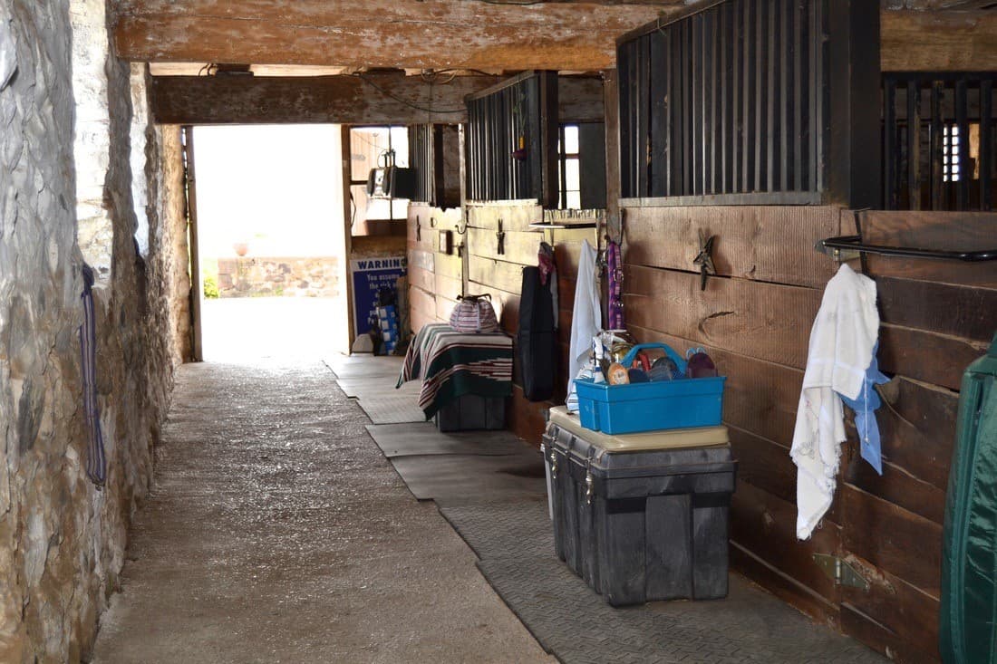 A rustic barn corridor with stables on either side and various supplies stored along the floor.