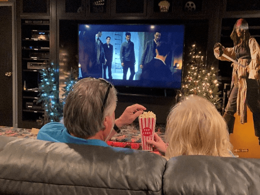 A couple enjoys a movie night with popcorn in a dimly lit room decorated with Christmas trees.