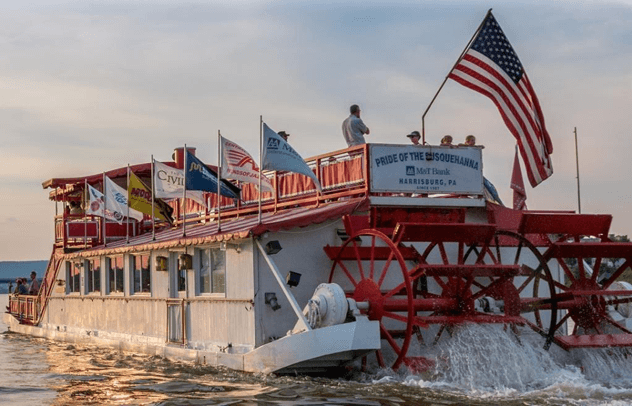 A red and white paddleboat, "Pride of the Susquehanna," cruises on a river, adorned with various flags and an American flag.