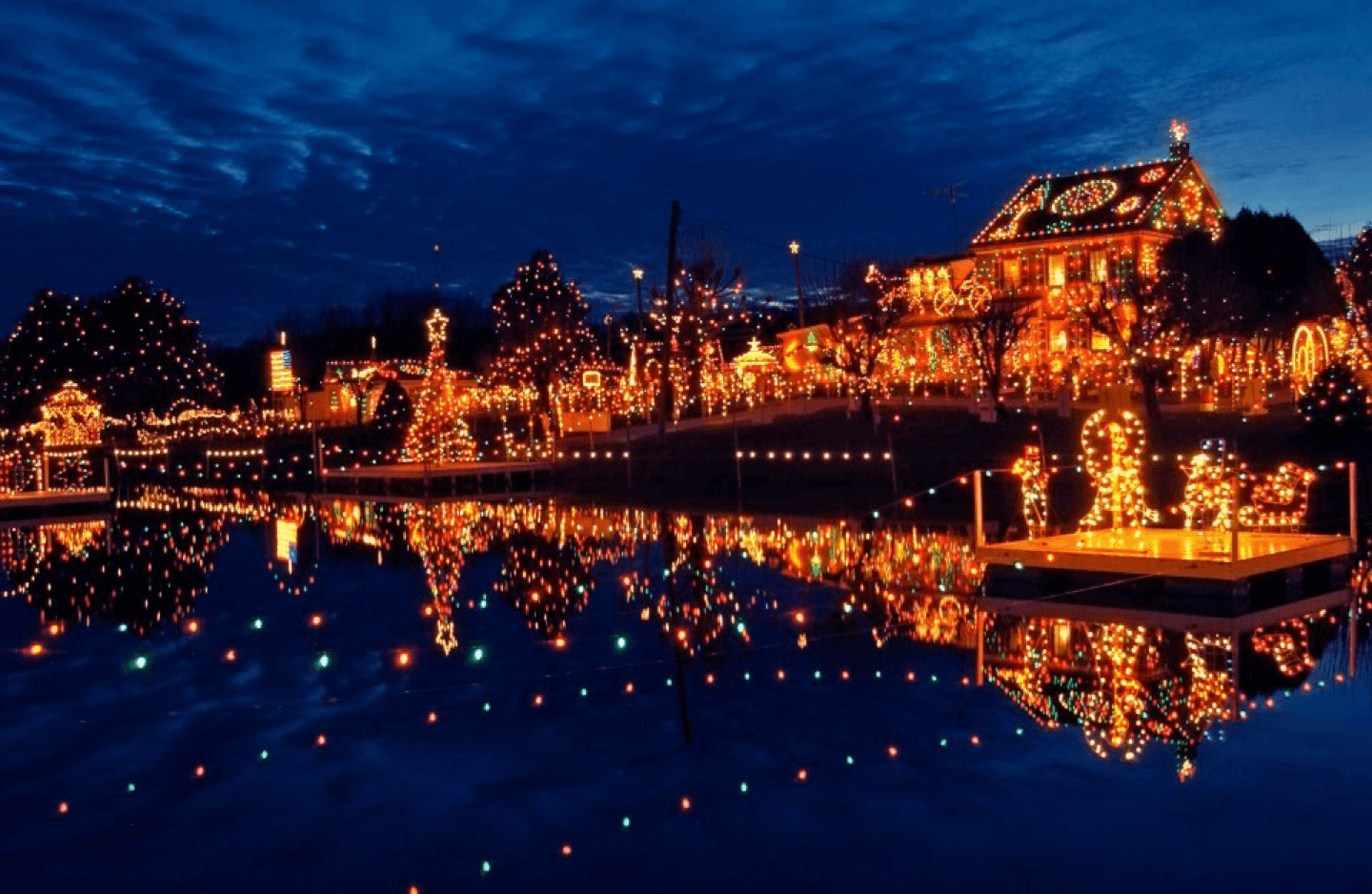A beautifully decorated house and garden with colorful Christmas lights reflecting in a calm lake under a dusk sky.