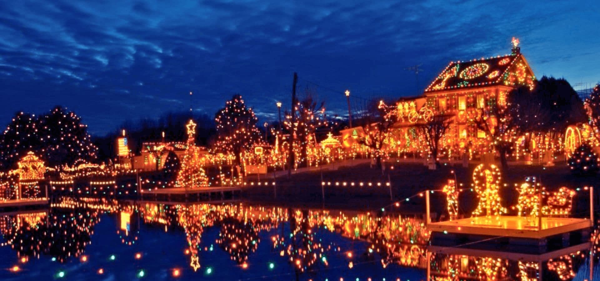 A beautifully illuminated house and landscape adorned with festive lights, reflecting in a calm water body under a twilight sky.