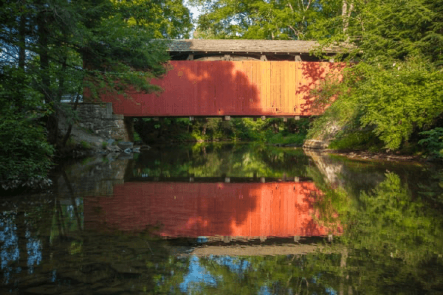A red covered bridge spans a peaceful stream surrounded by lush greenery.