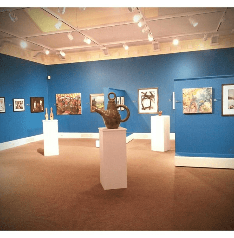 A vibrant art gallery featuring colorful paintings on blue walls and a central ceramic teapot sculpture.