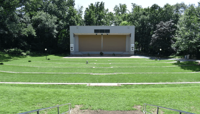 An outdoor amphitheater with a grassy seating area and a stage surrounded by trees.
