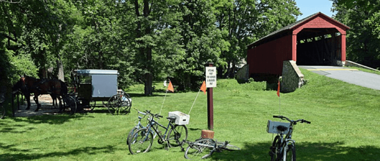Bicycles parked near a red covered bridge with horse-drawn carriages in a lush green setting.