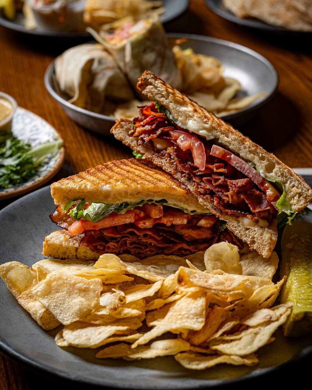 A stacked sandwich with bacon, lettuce, and tomatoes served alongside crispy potato chips on a black plate.