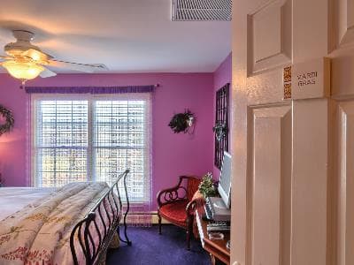 A cozy bedroom with pink walls, a metal bed frame, and a view out of bright windows, featuring a small seating area.