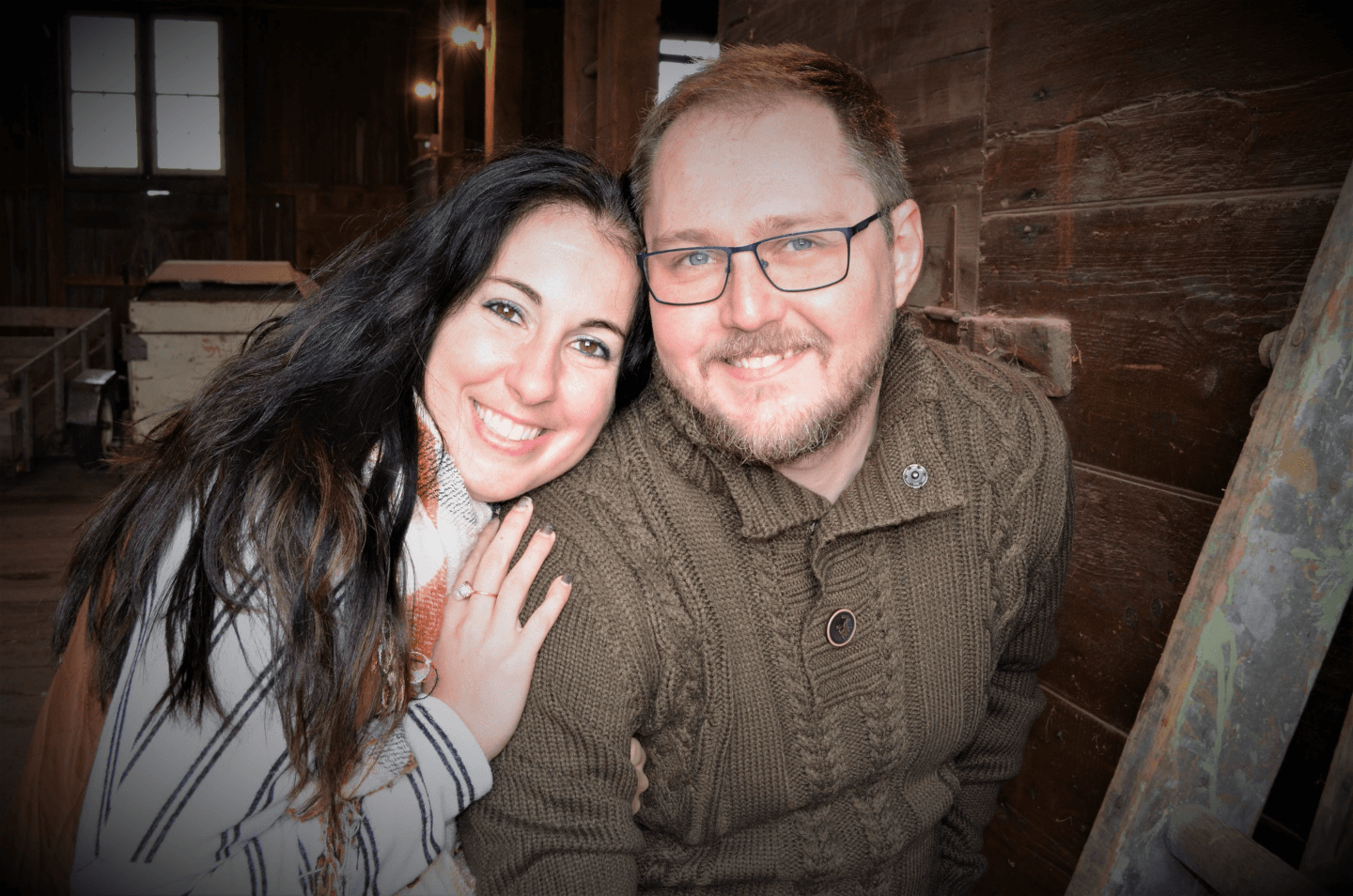 A smiling couple poses closely together in a rustic indoor setting.